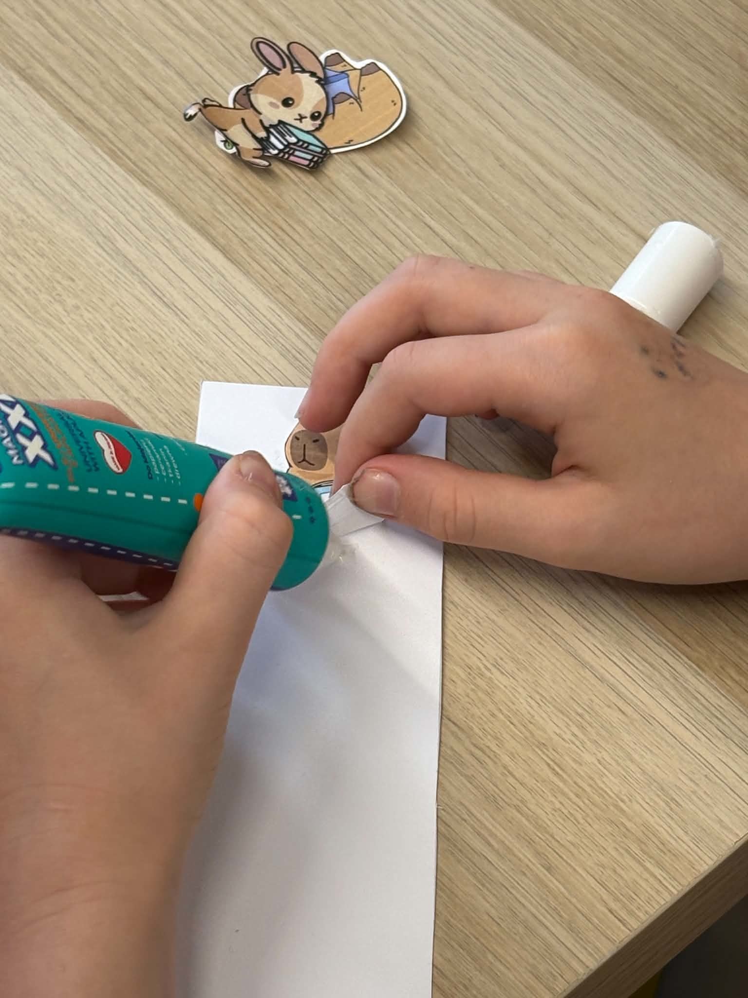 Zbliżenie na rączki dziecka przyklejającego papierową ilustrację kapibary do białego paska papieru za pomocą kleju w sztyfcie.