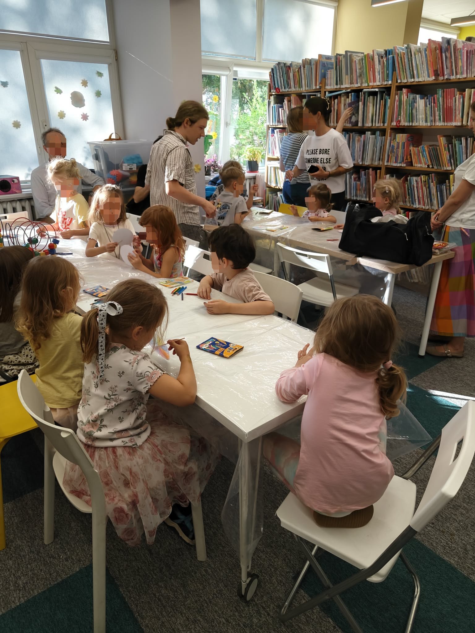 Ujęcie z boku na grupę dzieci siedzących przy długim stole w bibliotece. Na pierwszym planie dwie dziewczynki (w kwiecistej spódniczce i różowej bluzce) są skupione na kolorowaniu. W tle widać regały z książkami oraz dorosłych opiekunów obserwujących zajęcia. Jeden z instruktorów rozmawia z dzieckiem.