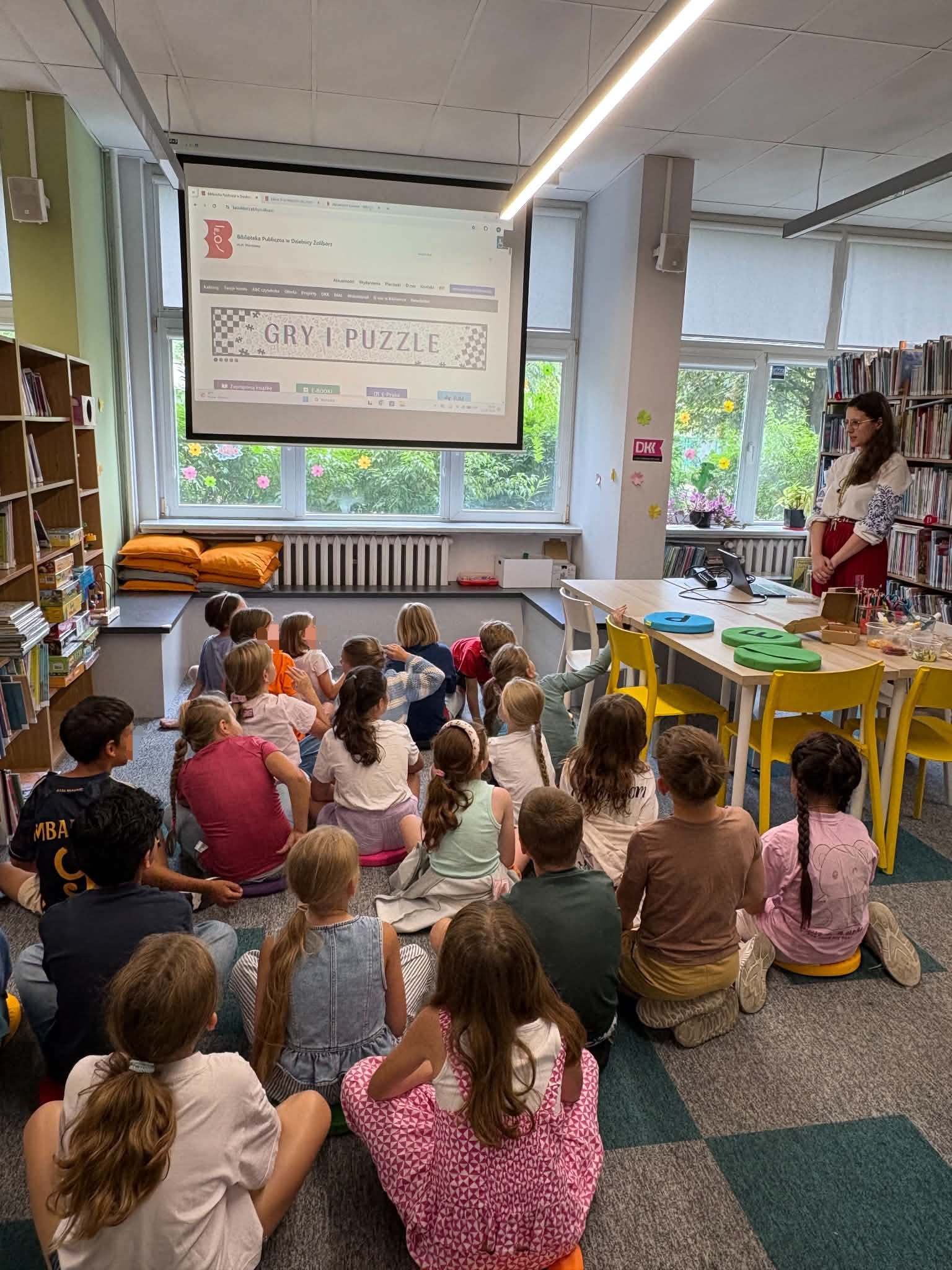 Dzieci siedzą na dywanie w bibliotece i patrzą na ekran projekcyjny, na którym wyświetlona jest strona internetowa z napisem „Gry i puzzle”. Obok ekranu stoi prowadząca w haftowanej bluzce.