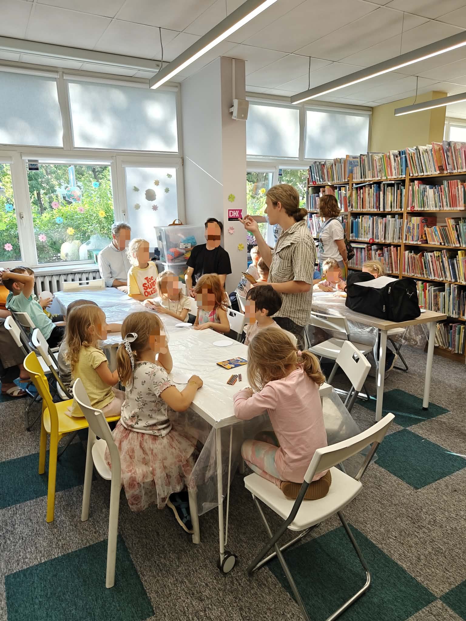 Szerokie ujęcie sali bibliotecznej wypełnionej dziećmi i dorosłymi podczas warsztatów. Dzieci siedzą przy dwóch stołach pod oknami. Pośrodku stoi instruktor (mężczyzna w jasnej koszuli), który gestykuluje ręką, tłumacząc coś grupie. Z prawej strony widać wysokie, pełne regały z książkami. Atmosfera jest jasna i twórcza.
