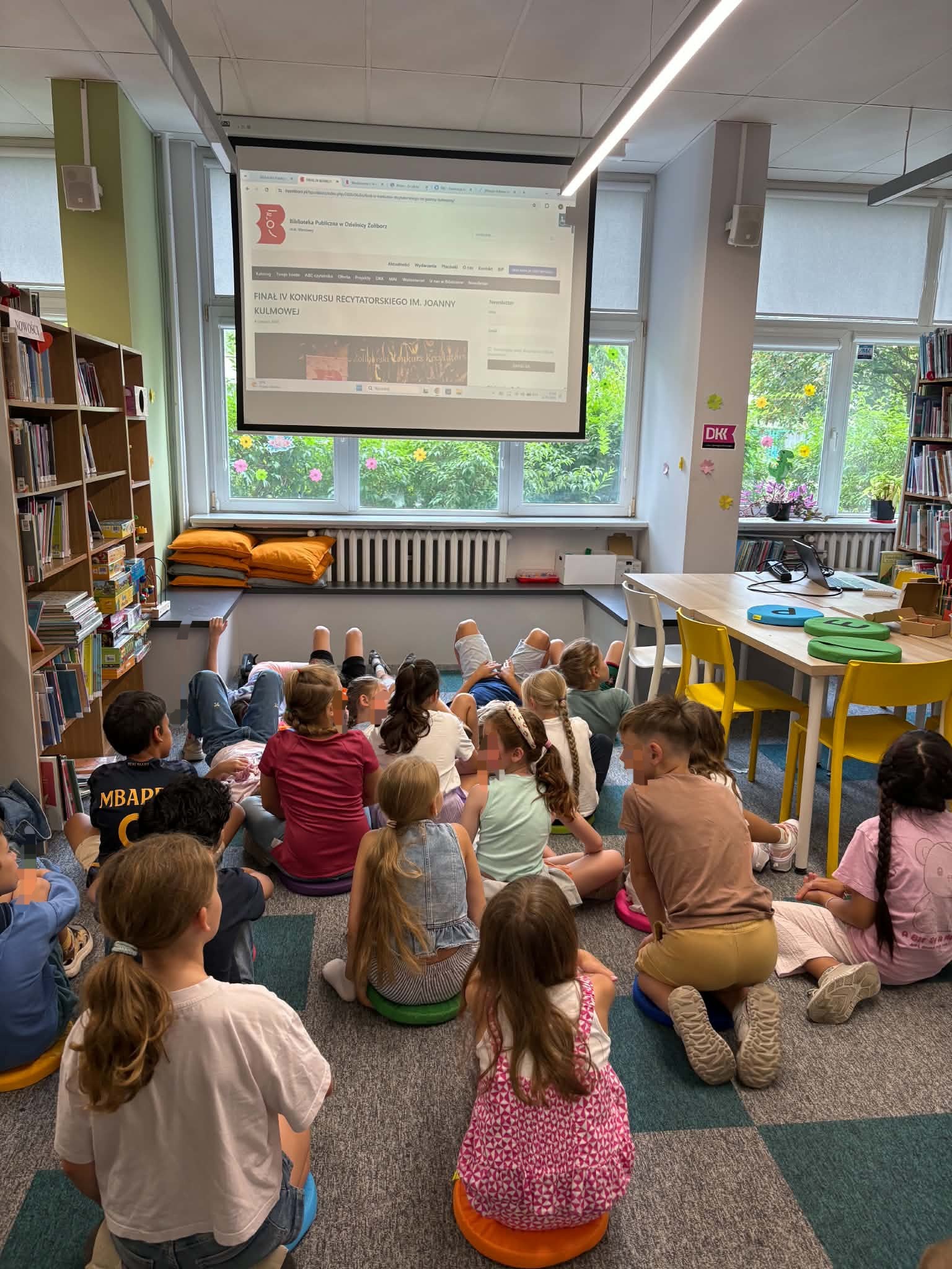 Dzieci siedzą i leżą na kolorowych poduszkach na dywanie w bibliotece, patrząc na ekran z wyświetloną stroną internetową informującą o finale konkursu recytatorskiego.