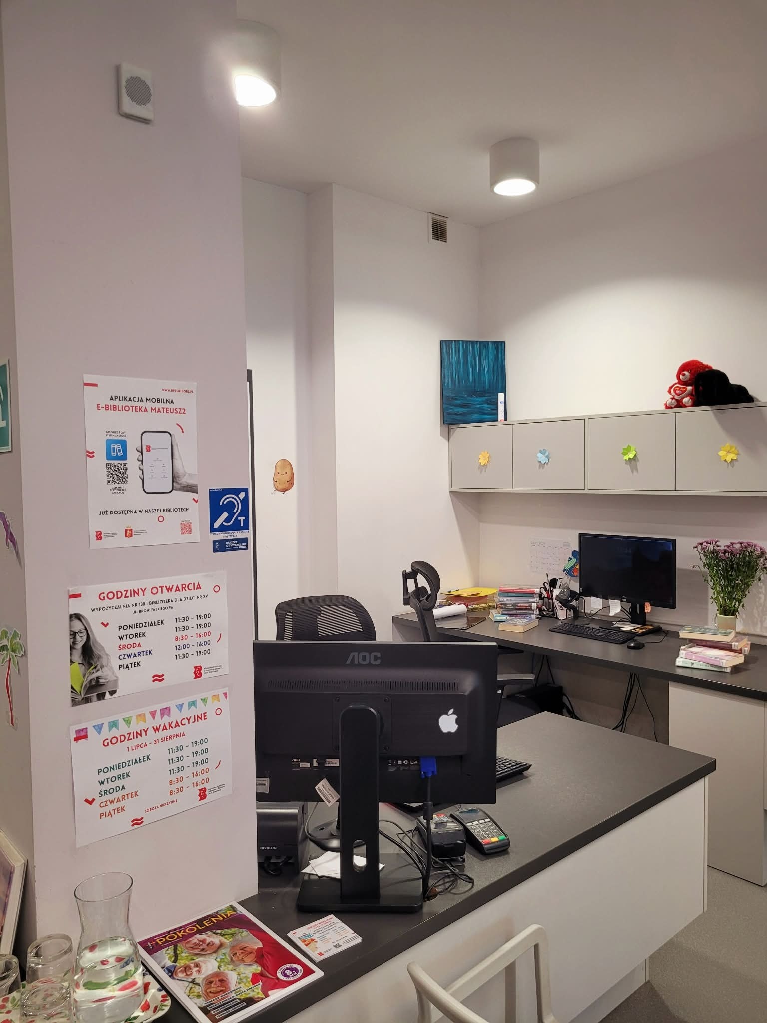 Stanowisko biblioteczne z ladą obsługi. Na ścianie obok znajdują się plakaty z godzinami otwarcia i oznaczenie dostępności, a wysoko przy suficie zamontowany jest znacznik systemu TOTUPOINT wspierający nawigację osób z dysfunkcją wzroku.