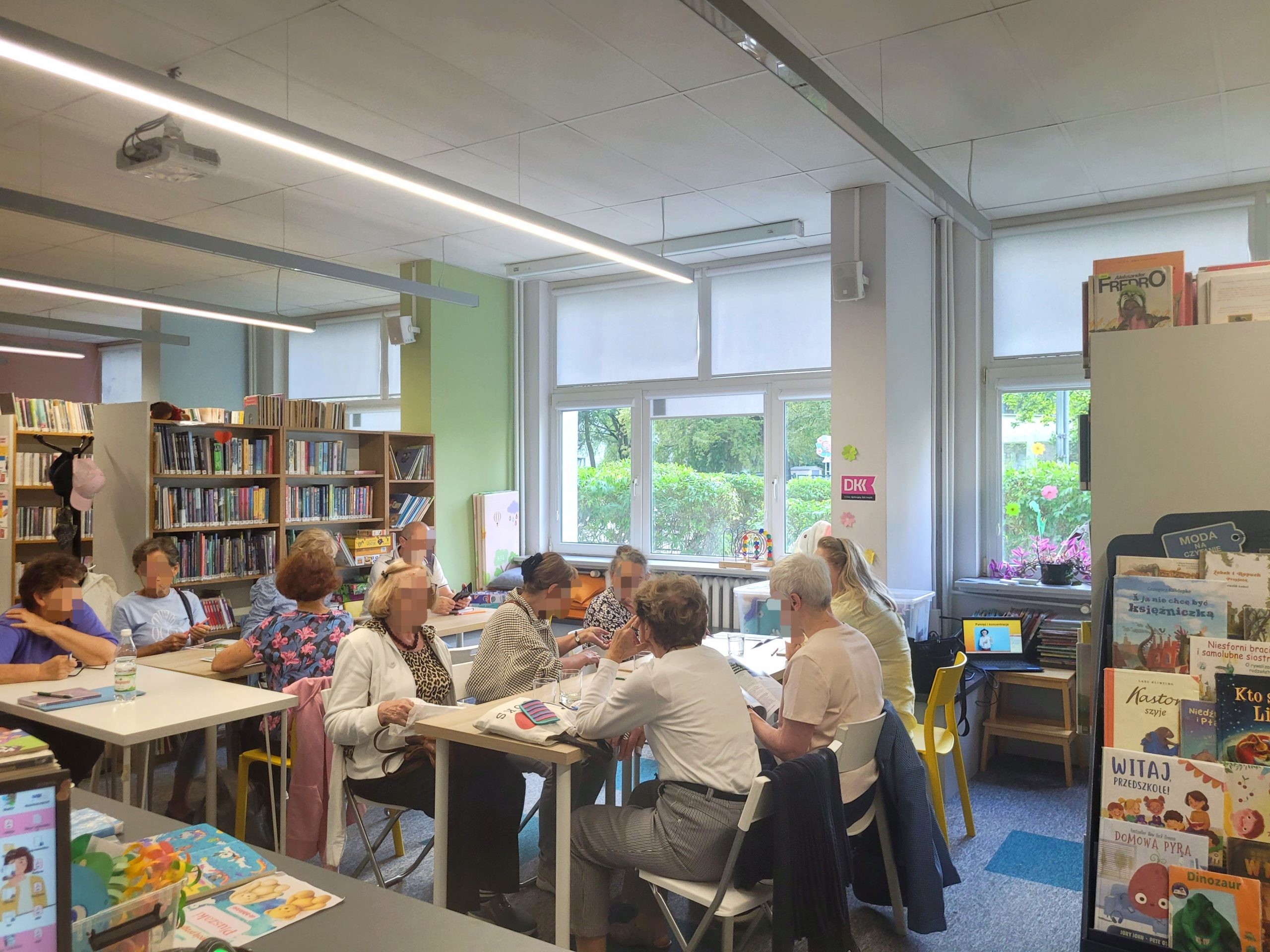 Widok na przestronną salę biblioteczną, w której przy ustawionych w rząd stołach siedzą osoby biorące udział w warsztatach lub spotkaniu. Po prawej stronie widać regał z ekspozycją książek dla dzieci.