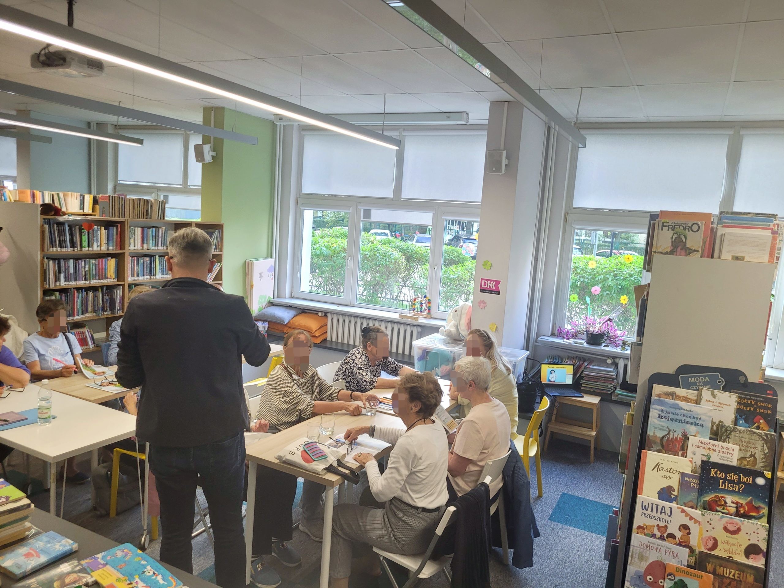 Prowadzący w ciemnym żakiecie stoi tyłem do obiektywu przed grupą seniorów pracujących przy stołach w jasnej bibliotece. Uczestnicy przeglądają drukowane arkusze.