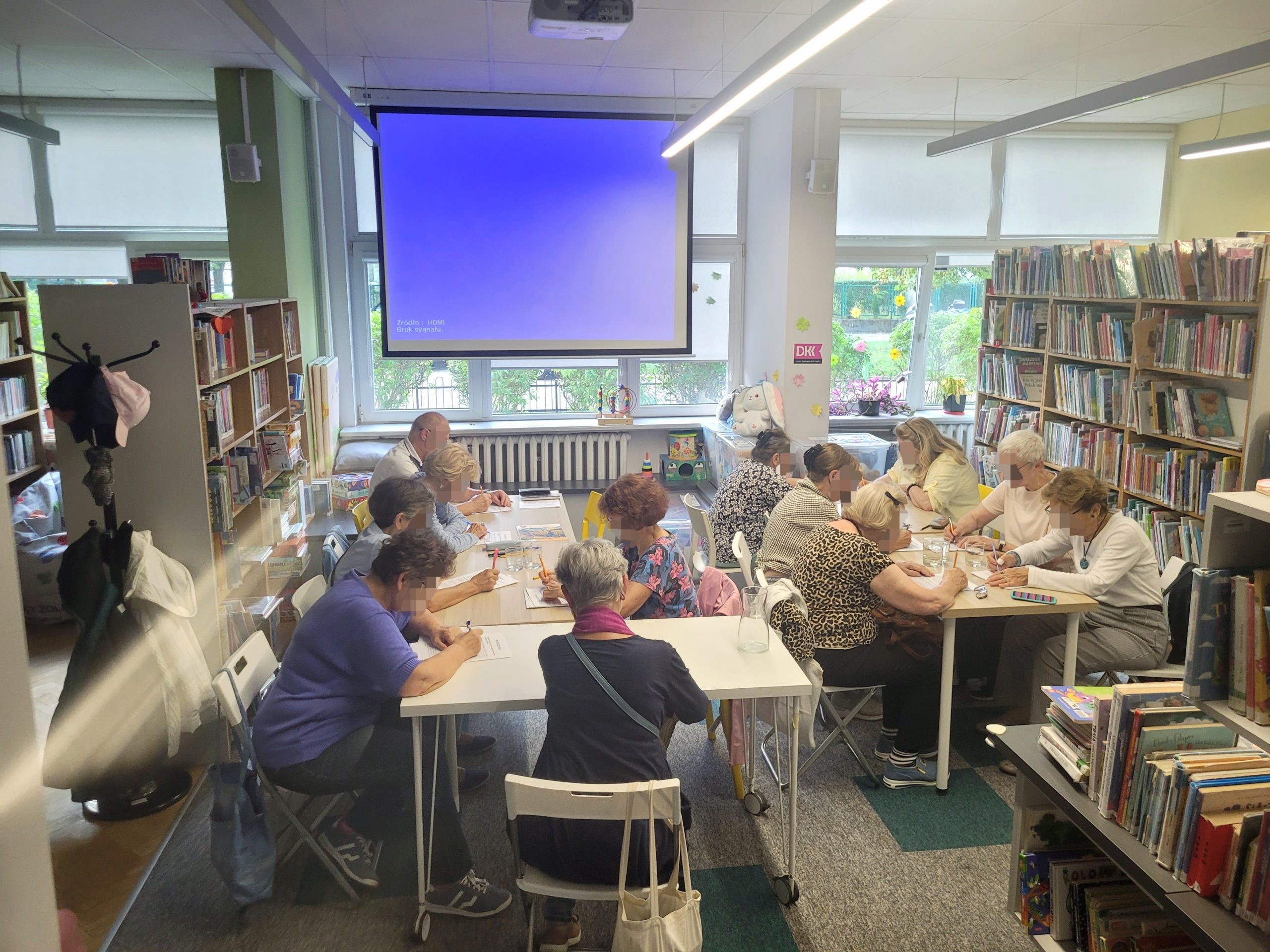 Szerokie ujęcie na salę biblioteczną z opuszczonym ekranem projekcyjnym. Uczestnicy spotkania siedzą przy stołach ustawionych w litery, pisząc i pracując nad arkuszami w otoczeniu regałów z książkami.