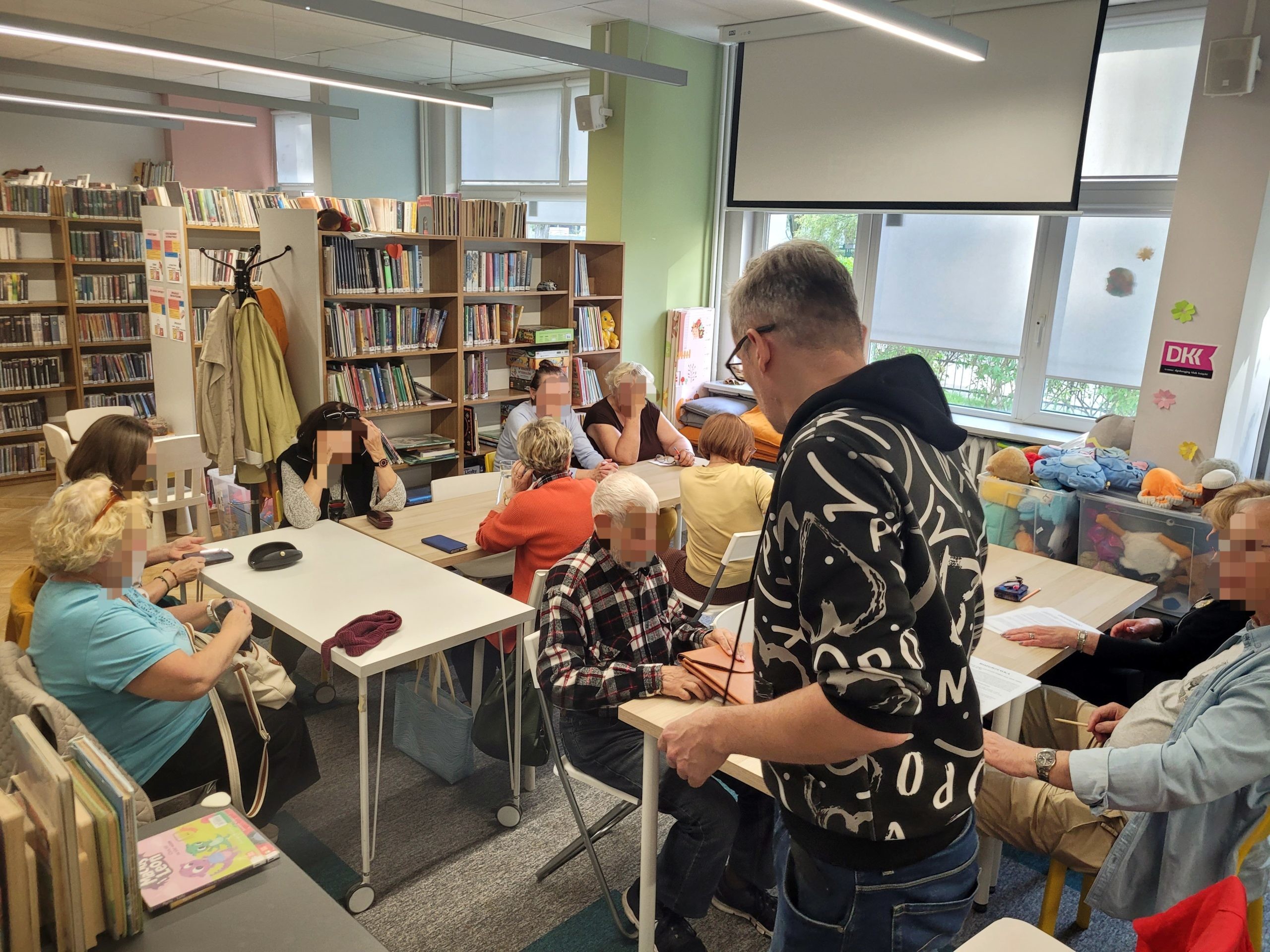 Grupa seniorek i seniorów siedzi przy stołach w bibliotece, wypełniając dokumenty lub rozwiązując zadania. Przed nimi stoi mężczyzna w czarnej bluzie we wzory, zorganizowany w dyskusję lub prowadzenie zajęć.