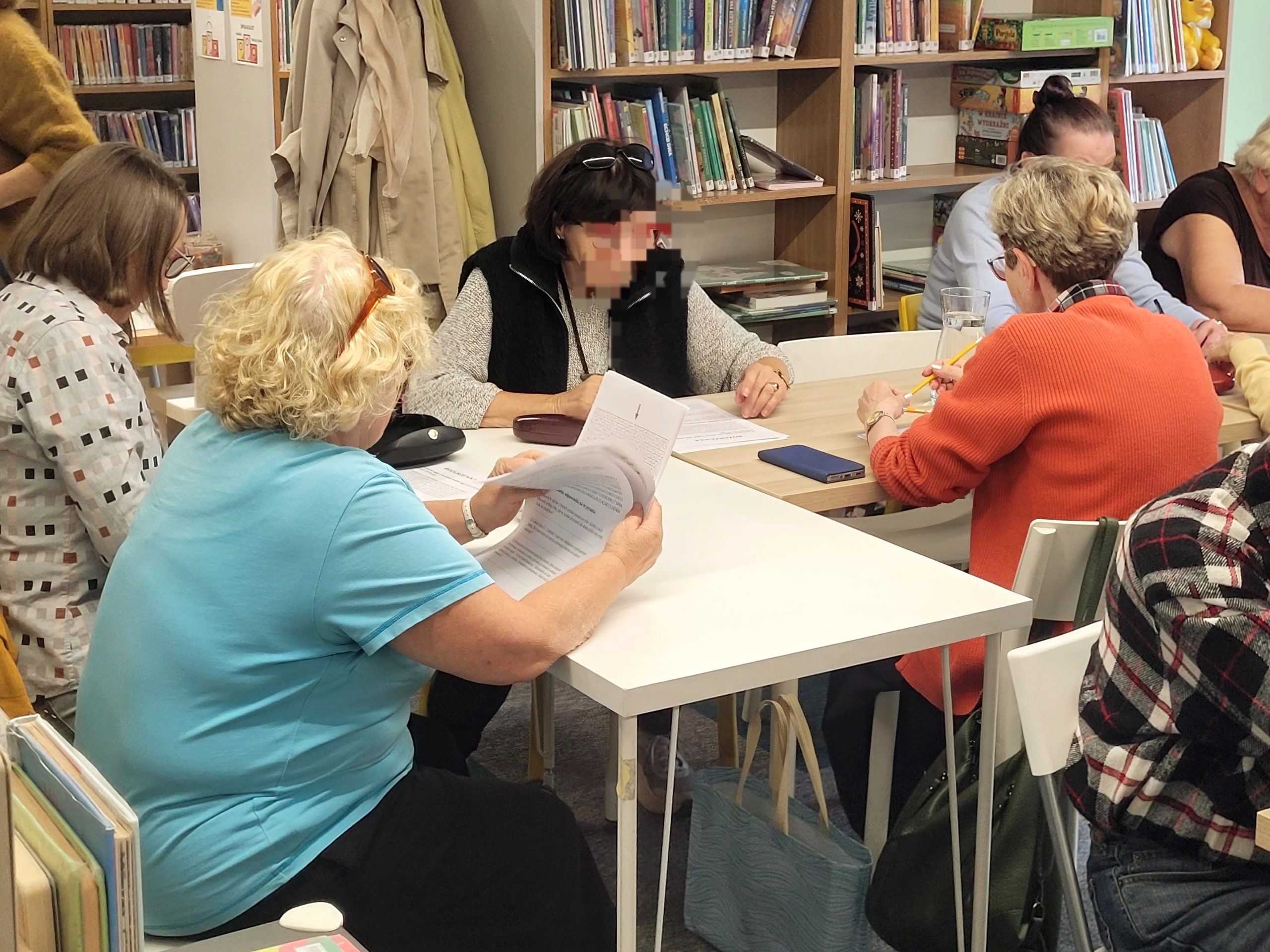Uczestniczki spotkania w bibliotece siedzą wokół złączonych stołów, przeglądają materiały papierowe i piszą długopisami. W tle widać regały pełne książek.