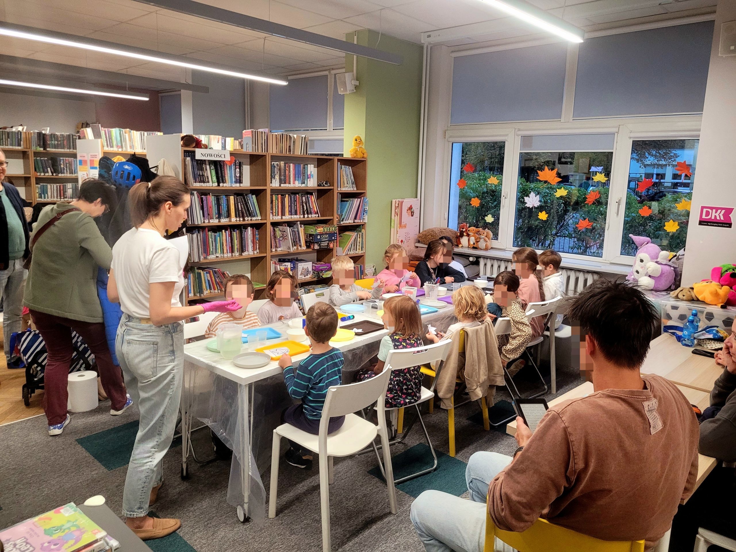 Szerokie ujęcie wnętrza jasnej biblioteki. Dzieci siedzą przy długim białym stole ustawionym na środku sali, zaangażowane w warsztaty plastyczne. Otaczają je wysokie regały wypełnione książkami, a na parapetach widać pluszowe zabawki i papierowe dekoracje w kształcie liści. Instruktorka w białym t-shircie stoi przy stole, a w tle widać inne osoby dorosłe. Przez duże okno wpada naturalne światło. Atmosfera jest przyjazna i twórcza. Twarze dzieci są zamazane, aby chronić ich prywatność.