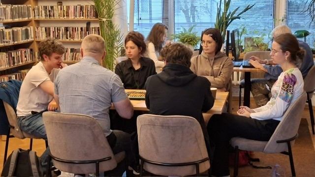 grafika wyróżniająca przedstawiająca uczestników Młodzieżowego Dyskusyjnego Klubu Książki (grupa starsza 16+) podczas omawiania książki "Myszy i ludzie". Sześciu uczestników siedzi wokół drewnianego stołu.