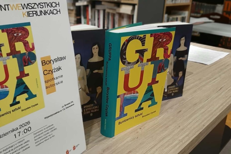 Grafika przedstawia zdjęcie ze spotkania autorskiego. Na drewnianym stole w bibliotece ustawiono egzemplarze książki „Gruppa. Buntownicy sztuki” Borysława Czyżaka oraz plakat zapowiadający spotkanie autorskie. Kolorowe okładki publikacji przyciągają uwagę na tle bibliotecznych regałów widocznych w głębi pomieszczenia, przygotowanego do rozmowy z autorem.