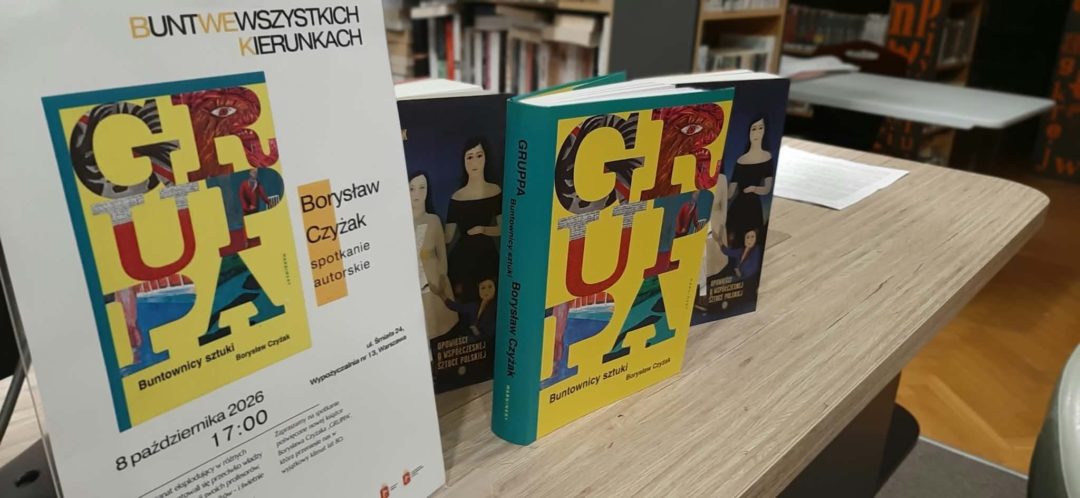 Grafika przedstawia zdjęcie ze spotkania autorskiego. Na drewnianym stole w bibliotece ustawiono egzemplarze książki „Gruppa. Buntownicy sztuki” Borysława Czyżaka oraz plakat zapowiadający spotkanie autorskie. Kolorowe okładki publikacji przyciągają uwagę na tle bibliotecznych regałów widocznych w głębi pomieszczenia, przygotowanego do rozmowy z autorem.